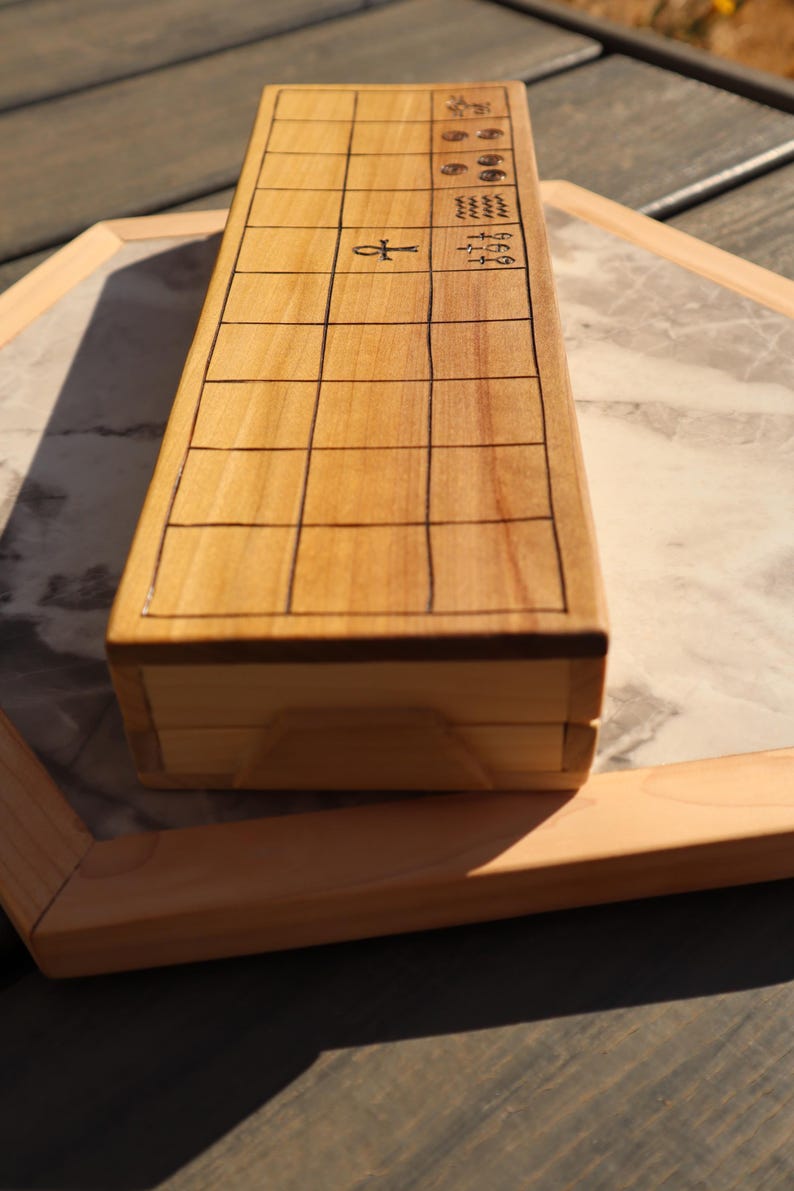 May include: A light-colored wooden box with a grid pattern on the top surface. The box has an ankh symbol and other hieroglyphic-like symbols. The box is on a marble-patterned surface.