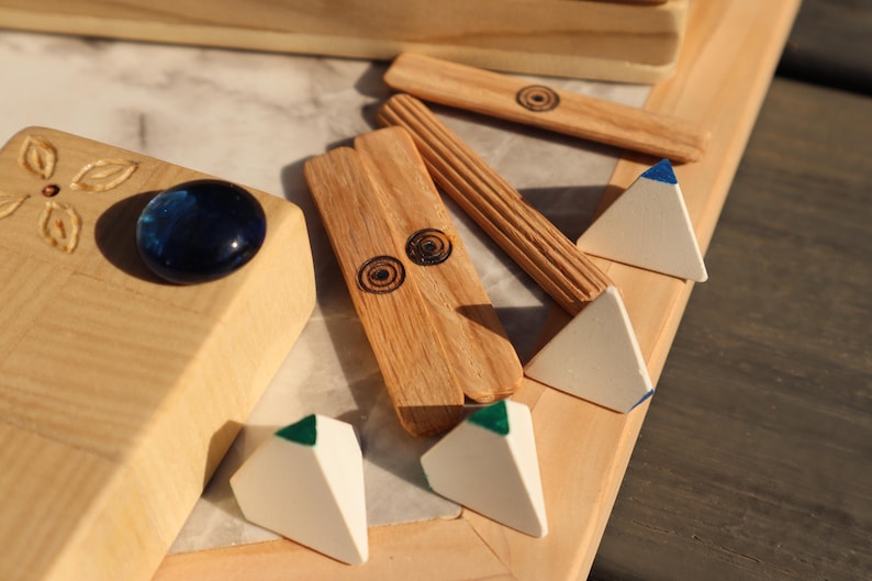 May include: Wooden game pieces and a decorative wooden box. The box has a blue glass stone and carved floral details. The game pieces include small white pyramids with colored tips and wooden sticks with etched designs.