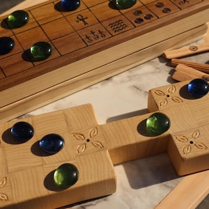 May include: Wooden board game with a light wood finish, featuring a grid pattern and decorative carvings. The game includes blue and green glass marbles, wooden game pieces, and a marble playing surface. The game is designed for two players.