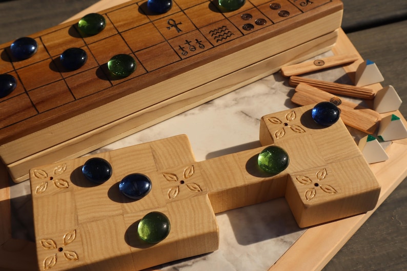 May include: Wooden board game with a light wood finish, featuring a grid pattern and decorative carvings. The game includes blue and green glass marbles, wooden game pieces, and a marble playing surface. The game is designed for two players.