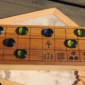 May include: A wooden board game with a grid pattern, featuring dark blue and green glass game pieces. The board has etched symbols and is set on a light-colored surface. A wooden frame is visible in the background.