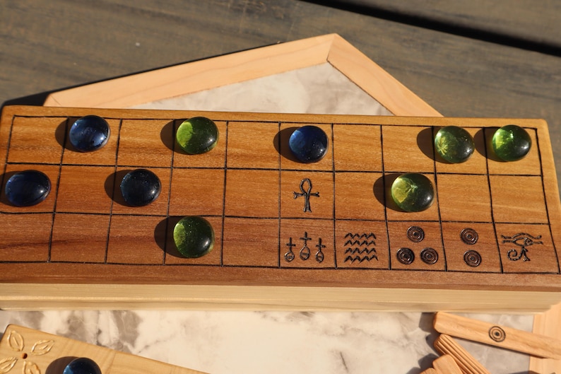 May include: A wooden board game with a grid pattern, featuring dark blue and green glass game pieces. The board has etched symbols and is set on a light-colored surface. A wooden frame is visible in the background.