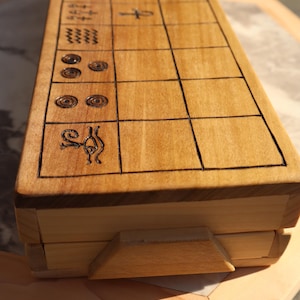 May include: A wooden game board with etched black lines and symbols. The board is rectangular with a light brown finish and a hinged lid. The game board is resting on a wooden surface.