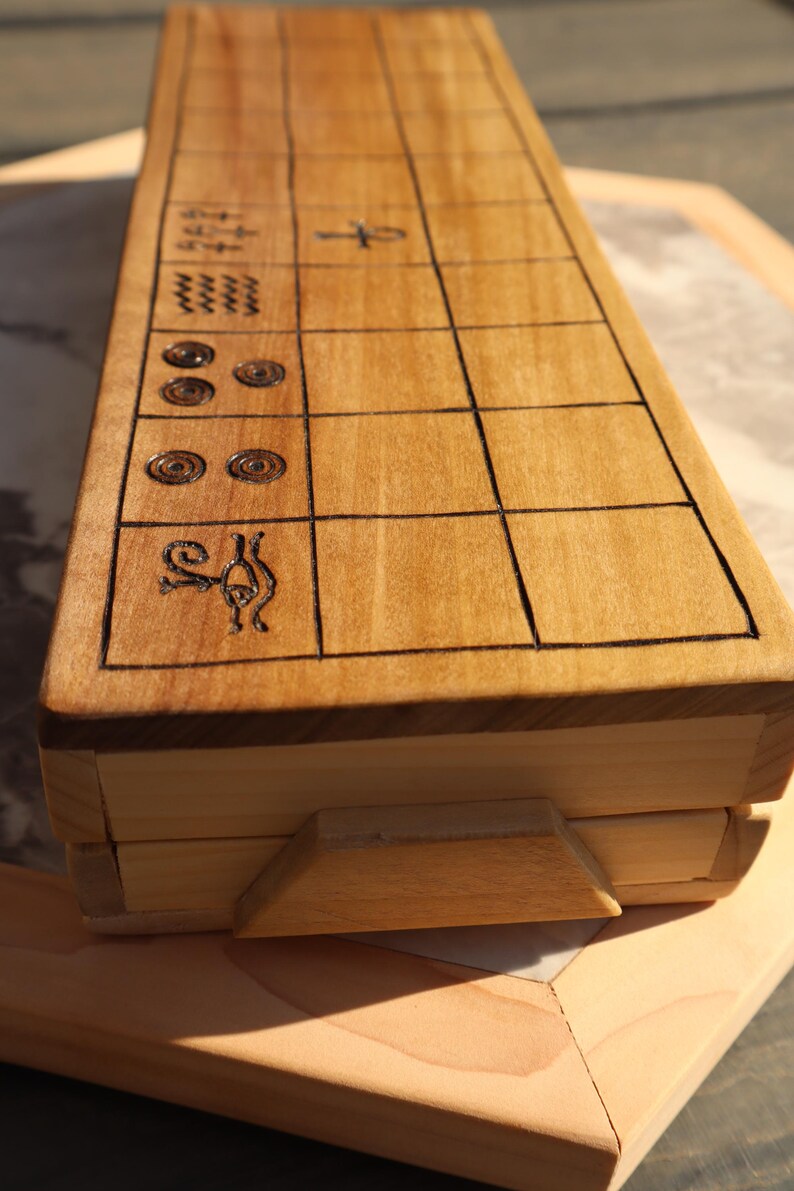 May include: A wooden game board with etched black lines and symbols. The board is rectangular with a light brown finish and a hinged lid. The game board is resting on a wooden surface.