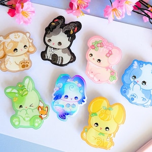 May include: A collection of seven acrylic pins featuring stylized cartoon rabbits in various colors and themes, including a beige rabbit with a bonnet, a black and grey rabbit, a pink strawberry-themed rabbit, a blue rabbit, a green rabbit with a fox, a blue and purple aquatic rabbit, and a yellow lemon-themed rabbit.