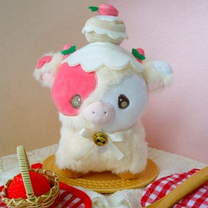 May include: A plush cow toy with a strawberry theme. The cow has a cream-colored body, pink patches, and a white frilly collar. A small strawberry and cream cake sits atop its head. A small basket of strawberries is in the foreground.