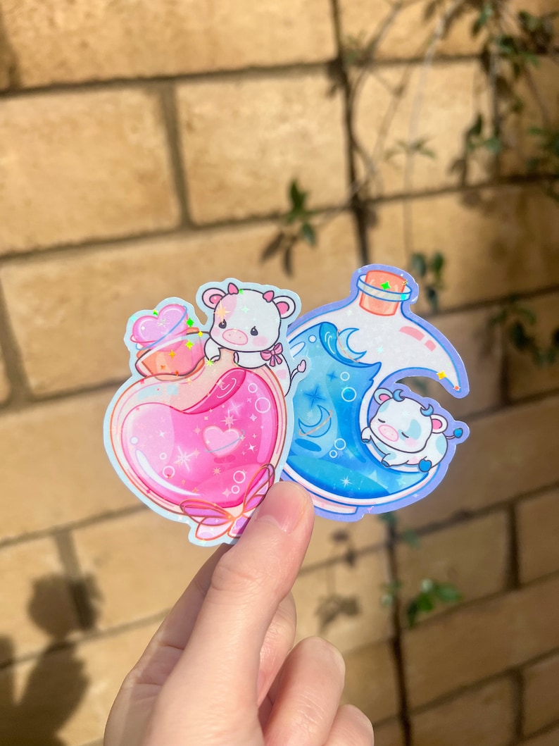 May include: Two stickers featuring a pink and blue bottle with a cow inside. The pink bottle has a heart and stars inside. The blue bottle has a moon and waves inside. Both bottles have a white cow with a pink bow.