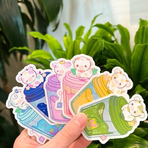 May include: Six colorful stickers featuring cartoon cows in glass jars with tea leaves. The stickers are arranged in a fan shape and are held in a hand.