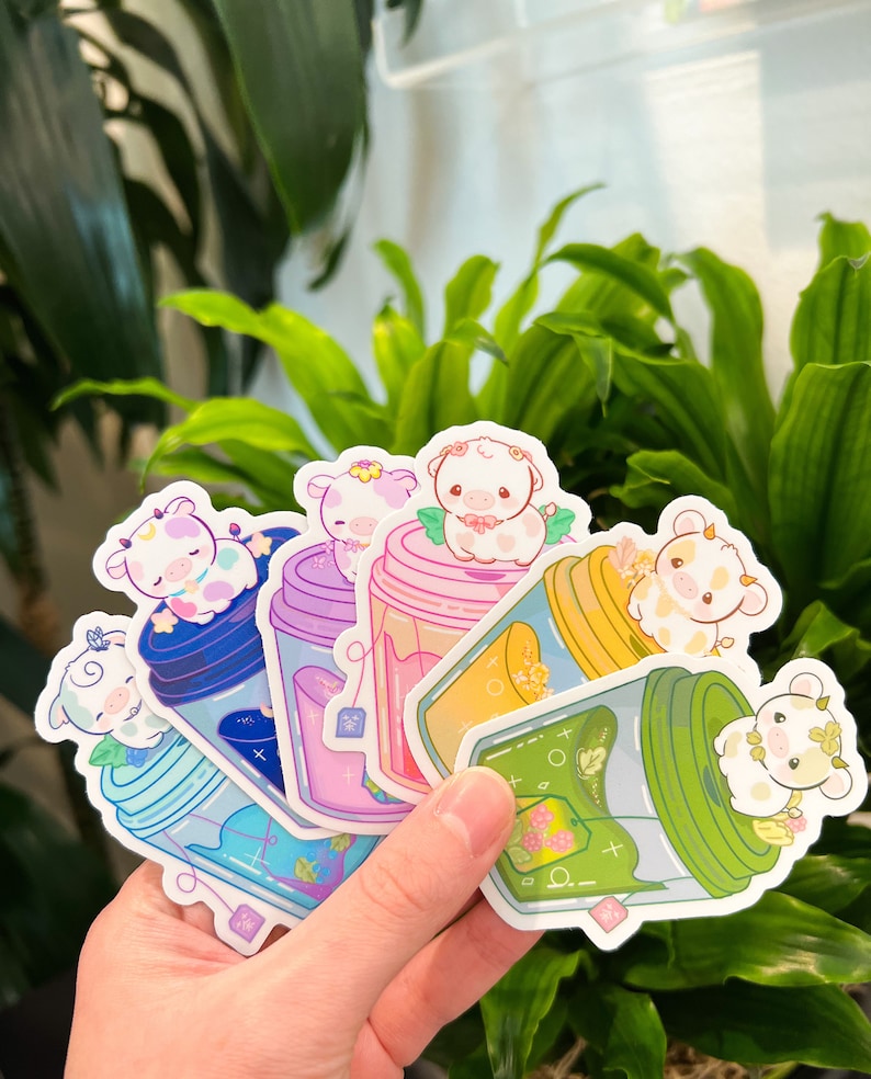 May include: Six colorful stickers featuring cartoon cows in glass jars with tea leaves. The stickers are arranged in a fan shape and are held in a hand.