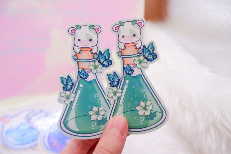 May include: Two white animal stickers with butterfly wings on a green potion bottle. The bottle is filled with a green liquid and has a white stopper. The animals are sitting on top of the stopper.