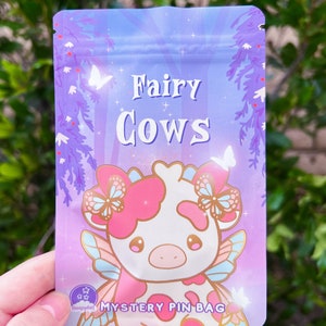 May include: A purple and pink holographic bag with a white cow with fairy wings. The bag says "Fairy Cows" and "Mystery Pin Bag".