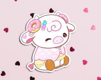 May include: A pink and white cartoon cow with a pink donut on its head. The cow is wearing a pink and white dress with hearts. The cow is sitting down and has a happy expression on its face.