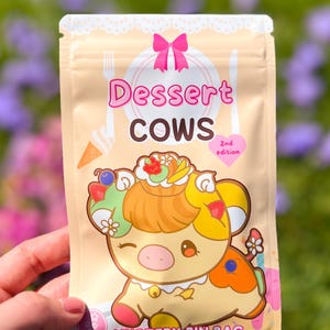May include: A tan-colored mystery pin bag featuring a cartoon cow with dessert-themed elements. The bag has the text "Dessert Cows" and "Mystery Pin Bag". The cow has a dessert-like design with a pink bow at the top.