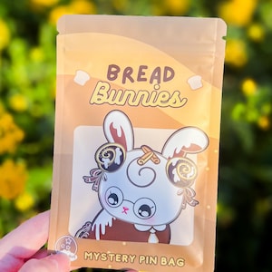 May include: A tan mystery pin bag with the words "Bread Bunnies" and a cartoon bunny illustration. The bunny has large eyes, glasses, and cinnamon roll-shaped ears. The bag also says "Mystery Pin Bag".