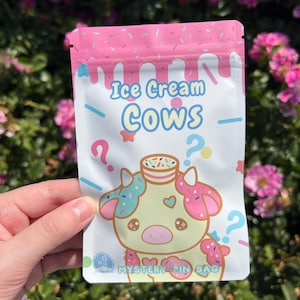 May include: A white and pink "Ice Cream Cows Mystery Pin Bag" is held in front of a green and pink floral background. The bag features a cartoon cow with ice cream-themed details and question marks. The top of the bag is pink with sprinkles.