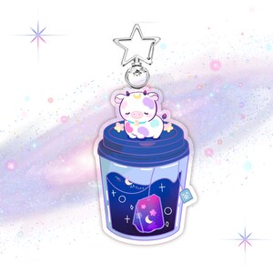 May include: A cute cartoon cow with white and pink spots is sleeping inside a blue and purple galaxy-themed teacup. The teacup has a tea bag with a moon and stars design. The teacup is attached to a silver star-shaped keychain.
