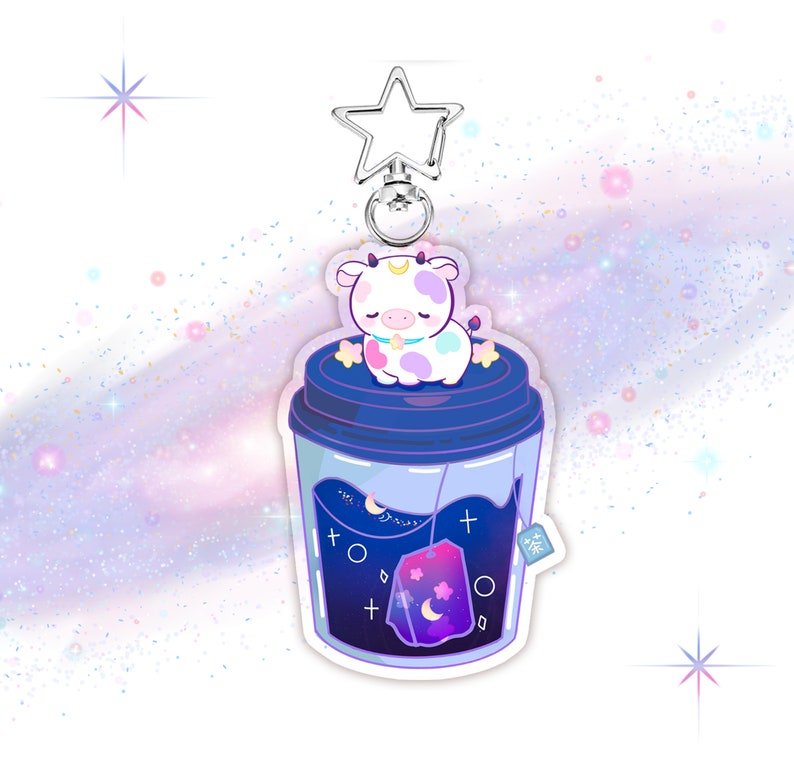 May include: A cute cartoon cow with white and pink spots is sleeping inside a blue and purple galaxy-themed teacup. The teacup has a tea bag with a moon and stars design. The teacup is attached to a silver star-shaped keychain.
