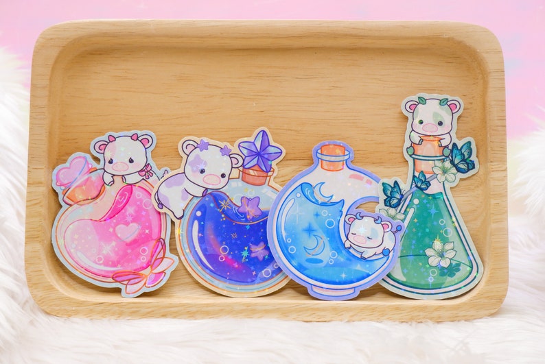 May include: Four colorful glass bottles with glitter and a cute cartoon cow inside each bottle. The bottles are on a wooden surface.