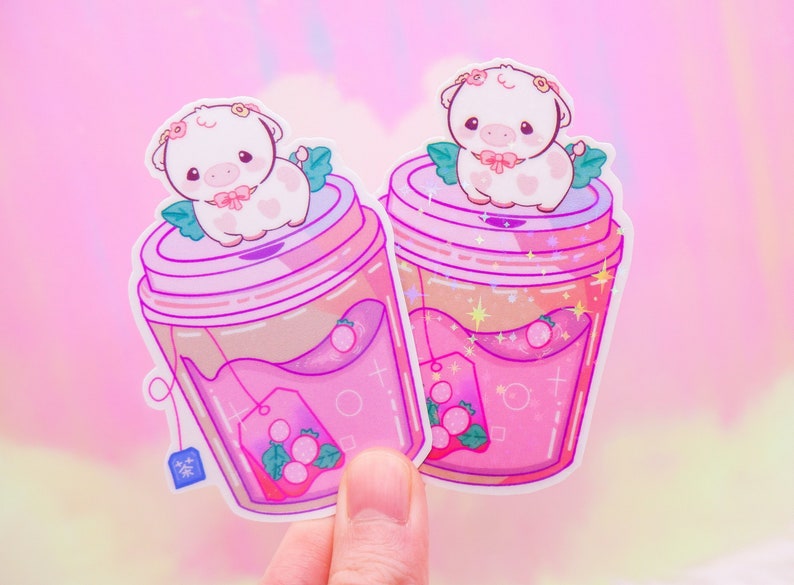 May include: Two pink and white stickers featuring a cartoon cow with a flower crown sitting on top of a pink and white cup of tea. The teacups have a pink and white tea bag with a green leaf and a pink and white flower.