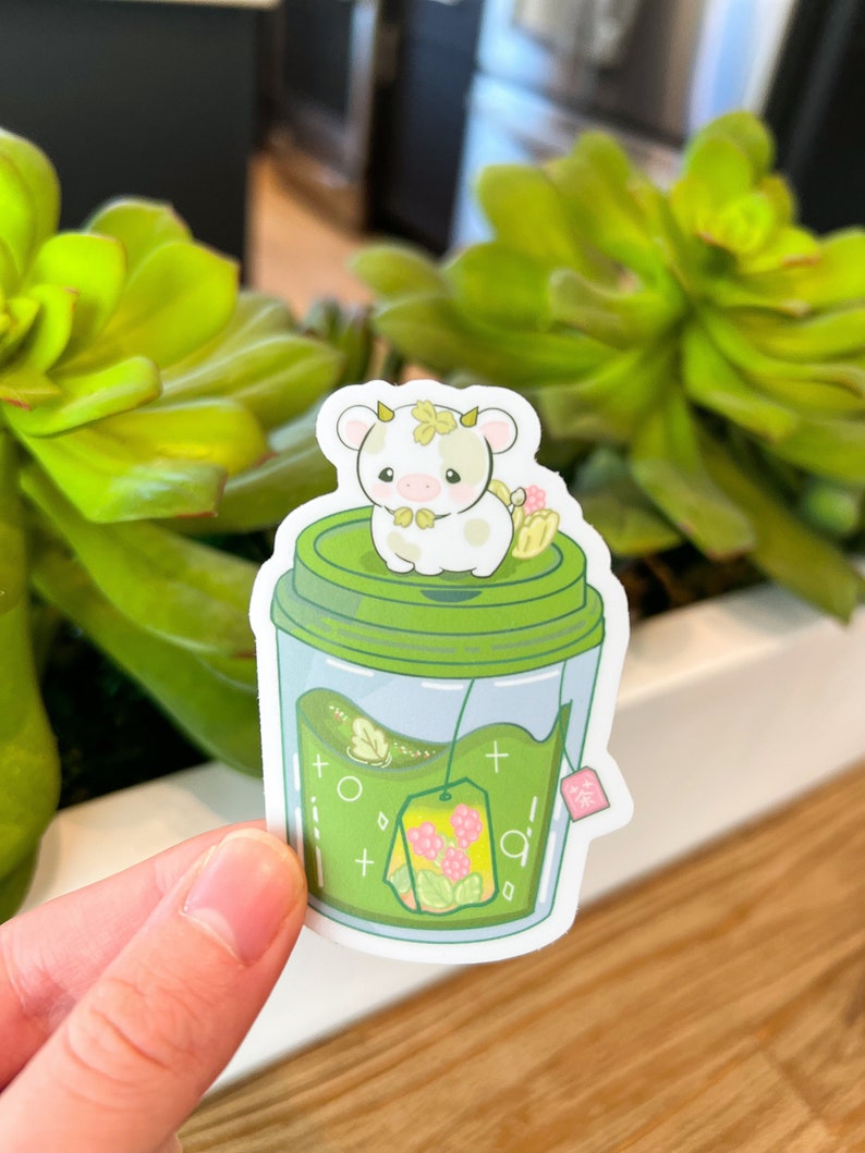 May include: A green and white sticker of a cartoon cow sitting on top of a green cup of tea with a tea bag. The tea bag has pink flowers and green leaves. The sticker has the text "茶" on the side.