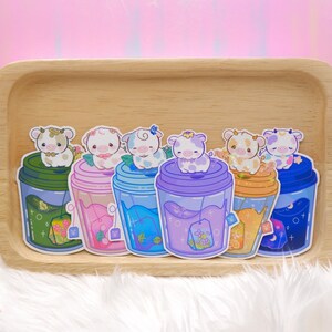 May include: Six colorful stickers featuring cartoon cows in cups of tea. Each cup has a different color and design, including green, pink, blue, purple, orange, and blue. The stickers are on a wooden tray.