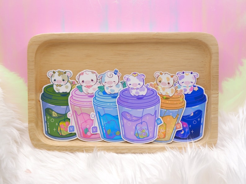 May include: Six colorful stickers featuring cartoon cows in cups of tea. Each cup has a different color and design, including green, pink, blue, purple, orange, and blue. The stickers are on a wooden tray.