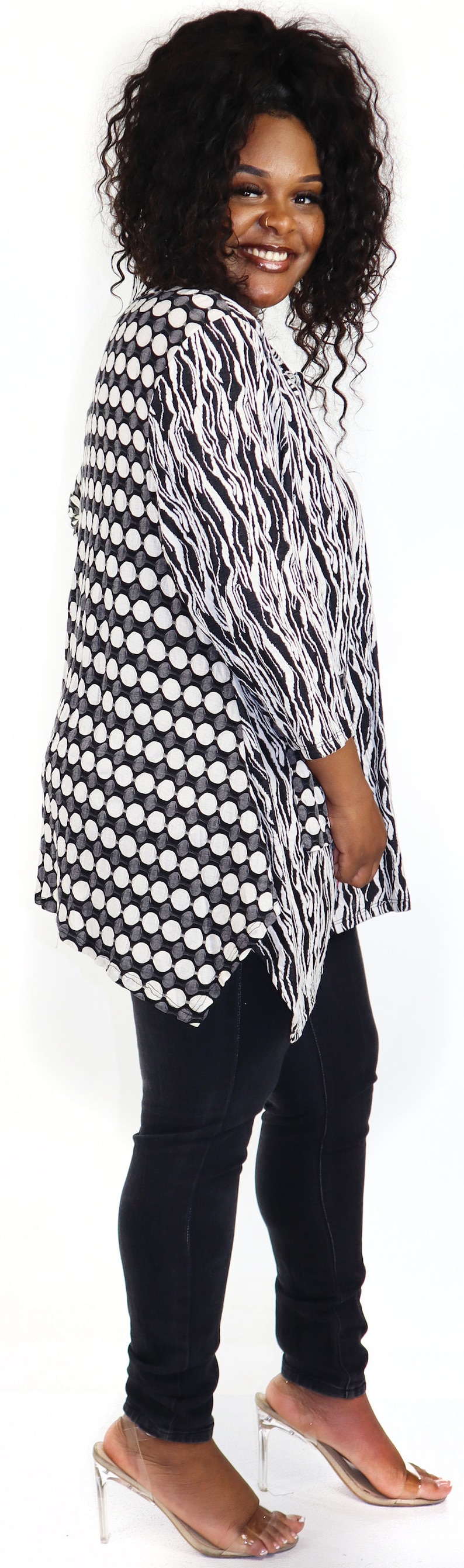 May include: A woman wearing a black and white patterned blouse with a black and white zebra print on one side and a black and white polka dot pattern on the other side. She is wearing black jeans and clear heeled sandals.