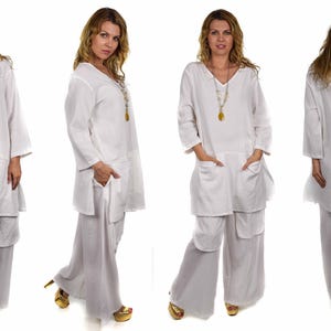 May include: A white linen tunic top with a v-neck and long sleeves. The top has two pockets on the front and a long, flowing design. The model is wearing the top with white linen pants.
