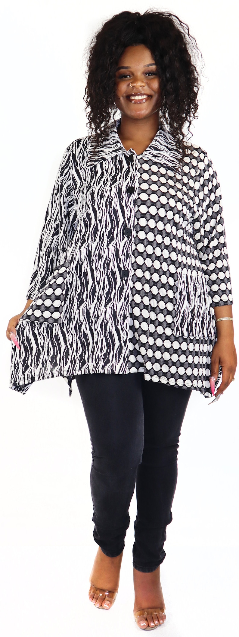 May include: A black and white patterned tunic with a long sleeve and a button-up front. The tunic has a wavy pattern on one side and a polka dot pattern on the other side. The model is wearing black leggings and clear heels.