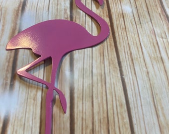 May include: Pink metal flamingo silhouette garden stake. The flamingo is standing on one leg with its head bent back. The stake is inserted into a wooden surface.