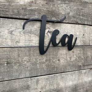 May include: Black metal sign with the word "Tea" written in a cursive font. The sign is mounted on a gray wooden wall.