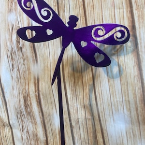 May include: A purple metal dragonfly garden stake. The dragonfly has heart-shaped cutouts and decorative swirls on its wings. The stake is designed to be placed in a garden or flower bed. The dragonfly is a decorative outdoor accent.