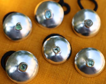 May include: Six circular hair ties featuring polished silver-toned dome-shaped ornaments. Each dome is centered with a small, square-cut, light green gemstone set in a gold-toned bezel. The hair ties are attached to black elastic bands.