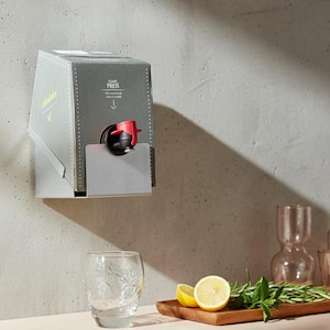Könnte beinhalten: Ein grauer Weinspender mit rotem Zapfhahn und silberner Tropfschale, an der Wand befestigt. Der Spender hat Text und einen Pfeil nach unten. Ein klares Glas, Zitronen und Kräuter liegen auf einem Holzbrett, mit Glasflaschen im Hintergrund.