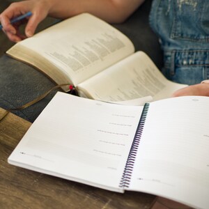 May include: An open spiral bound notebook with blank pages, lying on a wooden surface. A closed book with a brown leather cover is visible in the background.