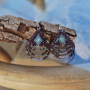 May include: Handmade beaded earrings with a geometric design. The earrings feature a triangular top with a central turquoise accent and cascading beaded strands in shades of burgundy, purple, and white. The earrings are suspended from silver-tone hooks.