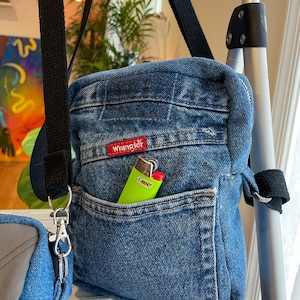 May include: A blue denim crossbody bag with a black strap and a zip closure. The bag has a large front pocket with the word "Wrangler" printed in red. A green lighter is in the pocket.
