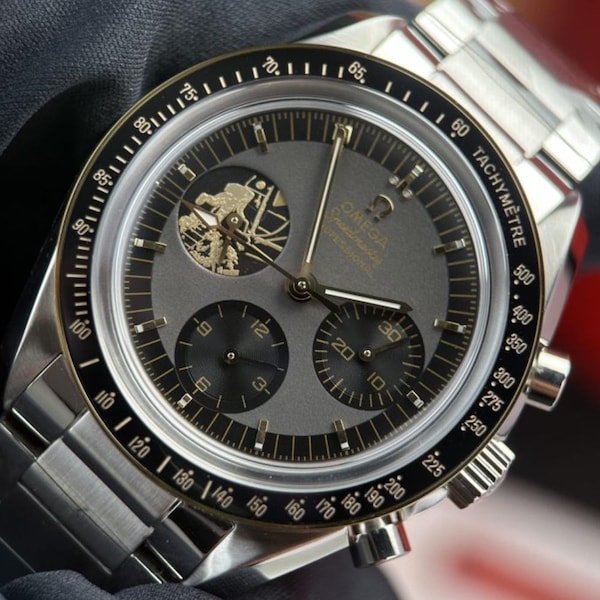 May include: An Omega Speedmaster Professional chronograph watch with a grey dial, three sub-dials, and a black tachymeter bezel. The watch features a stainless steel case and bracelet, with a detailed graphic of an astronaut on the sub-dial.