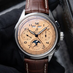 May include: A Breitling Datora watch with a copper-colored dial, featuring a moon phase display, chronograph sub-dials, day and month windows, and Arabic numerals. The timepiece has a silver-tone stainless steel case and a brown leather strap.