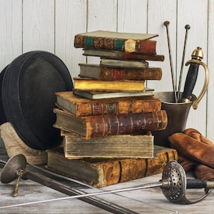 May include: A stack of antique leather-bound books positioned next to a black fencing mask, a metal cup containing two fencing foils, a pair of brown leather gloves, and a fencing foil with a perforated metal guard resting in the foreground.