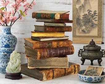 Op de afbeelding: Een stapel antieke, in leer gebonden boeken met verweerde ruggen en goudkleurige randen. De collectie bevat volumes met verschillende texturen en kleuren, rustend op een oppervlak naast een blauw-witte keramische vaas, een jadekleurig snijwerk, een bronzen wierookbrander en een kleine kom.