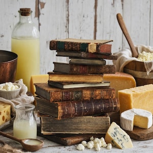 Op de afbeelding: Een stapel antieke, verweerde boeken met harde kaft, omringd door diverse kazen, waaronder een stuk blauwe kaas, een ronde brie en blokken gele kaas. Een glazen fles met vloeistof, een kommetje met wrongel en een houten lepel zijn aanwezig.