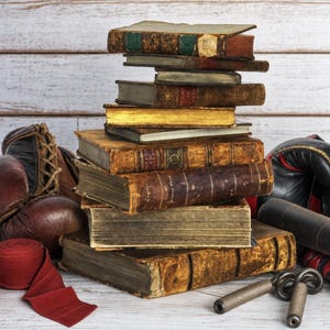 May include: A vertical stack of ten antique, weathered hardcover books with worn brown, gold, and dark green spines. Flanking the books are a pair of vintage brown leather boxing gloves, a red hand wrap, and a pair of black boxing gloves.