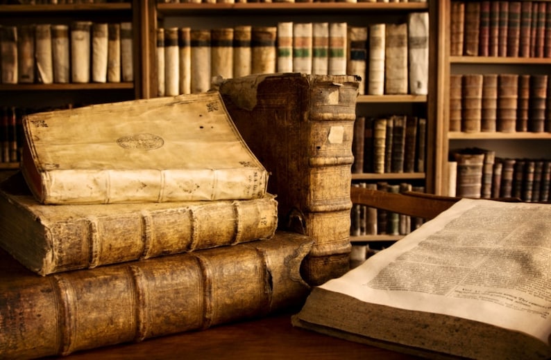 K&ouml;nnte beinhalten: Ein Stapel antiker, ledergebundener B&uuml;cher mit abgenutzten Seiten und goldenen Akzenten, der auf einem Holztisch vor einem B&uuml;cherregal mit weiteren B&uuml;chern steht.
