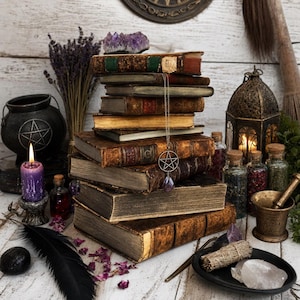 Pode incluir: Uma pilha de livros vintage desgastados com um aglomerado de ametista no topo, apresentando um colar com pingente de pentáculo de prata sobre as lombadas. A cena inclui um caldeirão preto, uma vela roxa, uma lanterna, frascos e um almofariz.