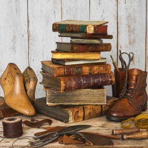 May include: A stack of antique leather-bound books sits beside a wooden shoe last, a brown leather lace-up boot, a spool of dark thread, metal shears, and a yellow measuring tape on a wooden surface with scattered leather scraps.