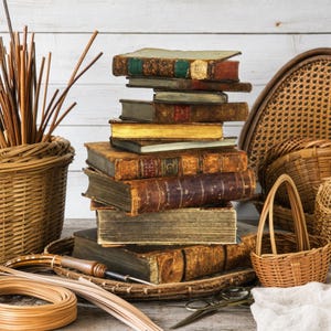 May include: A stack of antique books with worn leather covers and gold-gilded edges, surrounded by various woven wicker baskets, bundles of thin wooden reeds, a metal awl tool, and a pair of metal scissors on a wooden surface.