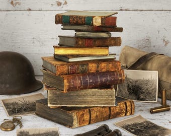 May include: A stack of antique books with worn leather covers, a dark brown military helmet, vintage binoculars, a canvas bag, and several black and white photographs depicting historical scenes on a light wooden surface.