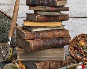 May include: A stack of nine weathered, leather-bound antique books with worn spines and edges. Beside the books are a fishing rod with a metal reel, a woven wicker basket containing colorful fishing lures, and a smooth gray stone with two flies.