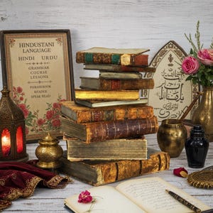 May include: A stack of antique books with worn leather covers and gold-gilded edges sits beside an open journal, a brass lantern, a metal vase with pink roses, and a framed sign titled Hindustani Language with Hindi and Urdu grammar lessons.